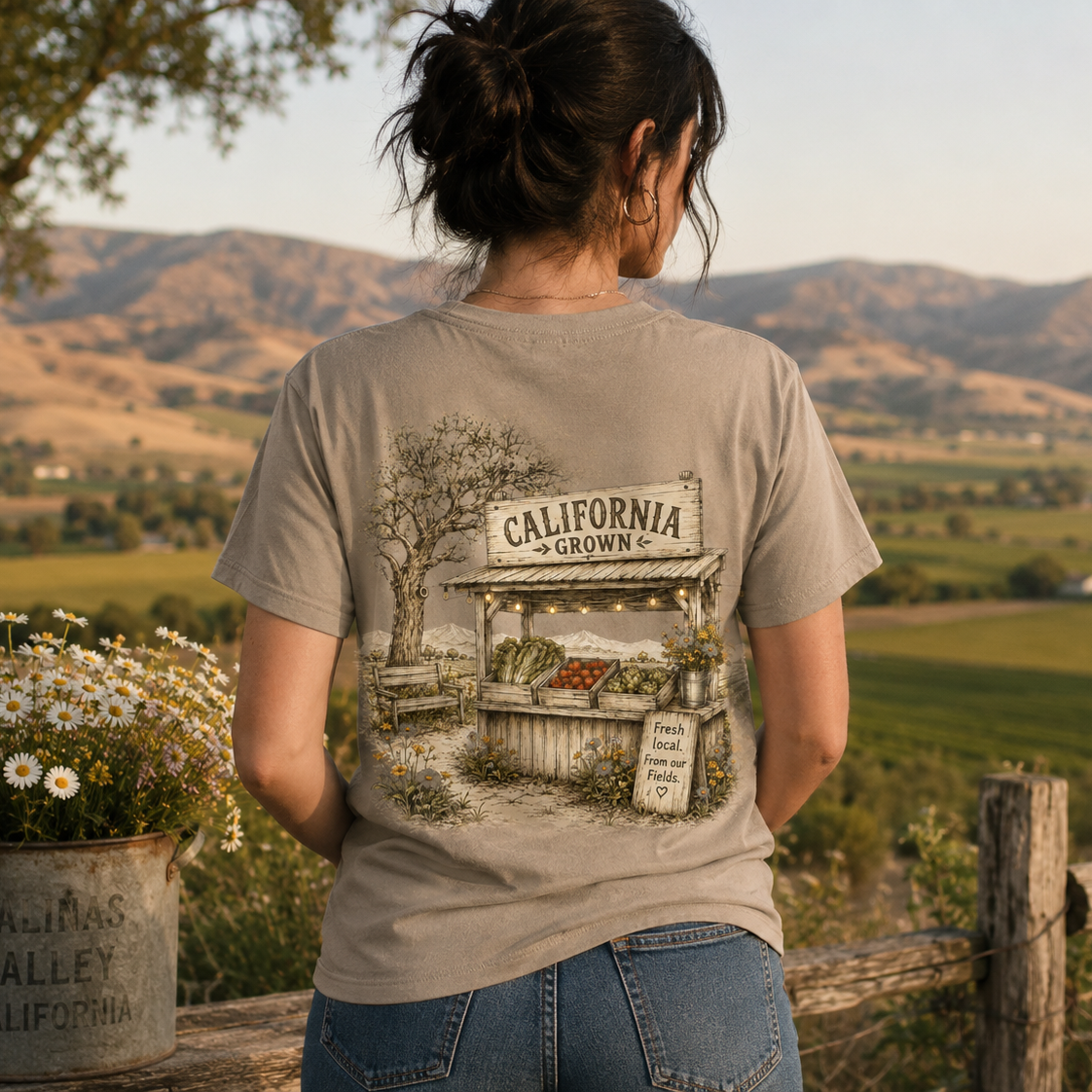 A woman viewed from behind wearing a beige graphic t-shirt with a vintage-style illustration of a 'California Grown' fruit stand, set against a scenic rural landscape with rolling hills and a flower pot in the foreground.