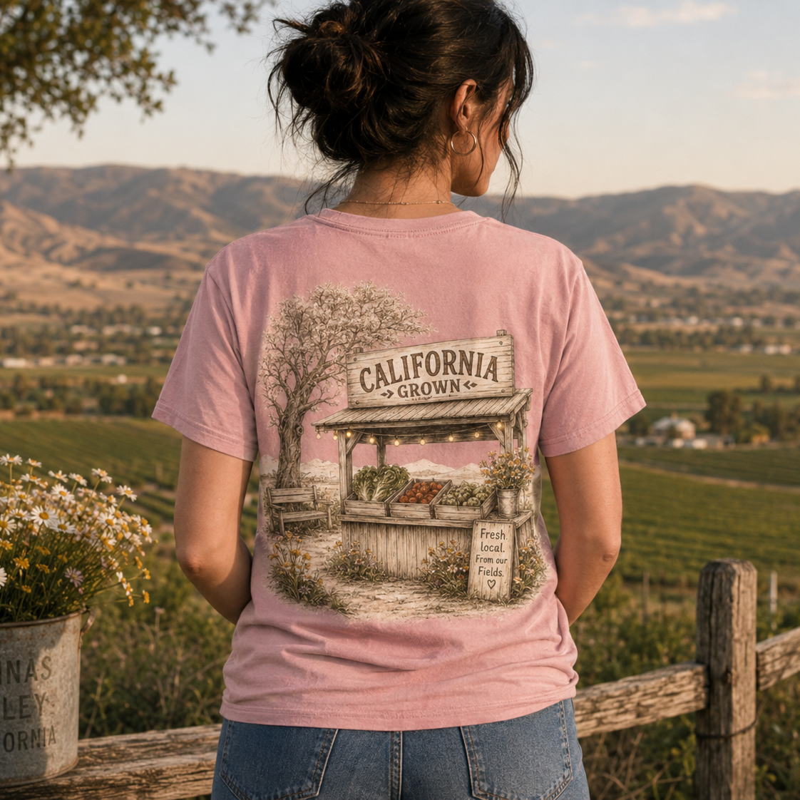 A woman viewed from behind wearing a light pink graphic t-shirt with a vintage-style illustration of a 'California Grown' orchid stand. The background shows a scenic vineyard landscape with rolling hills under a warm sky.