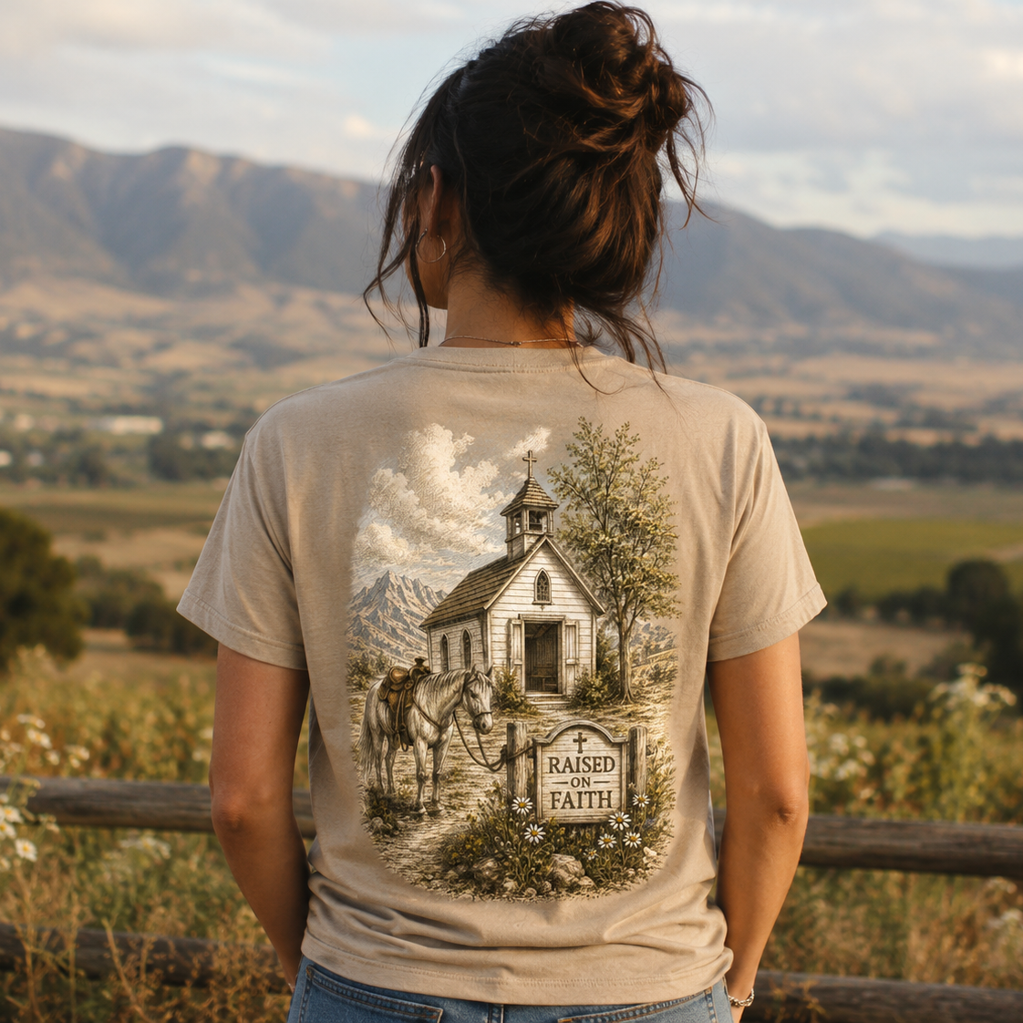 A woman viewed from behind wearing a beige graphic t-shirt with a detailed illustration of a church, horse, and a sign reading 'RAISED IN FAITH', set against a scenic outdoor background of mountains and fields.