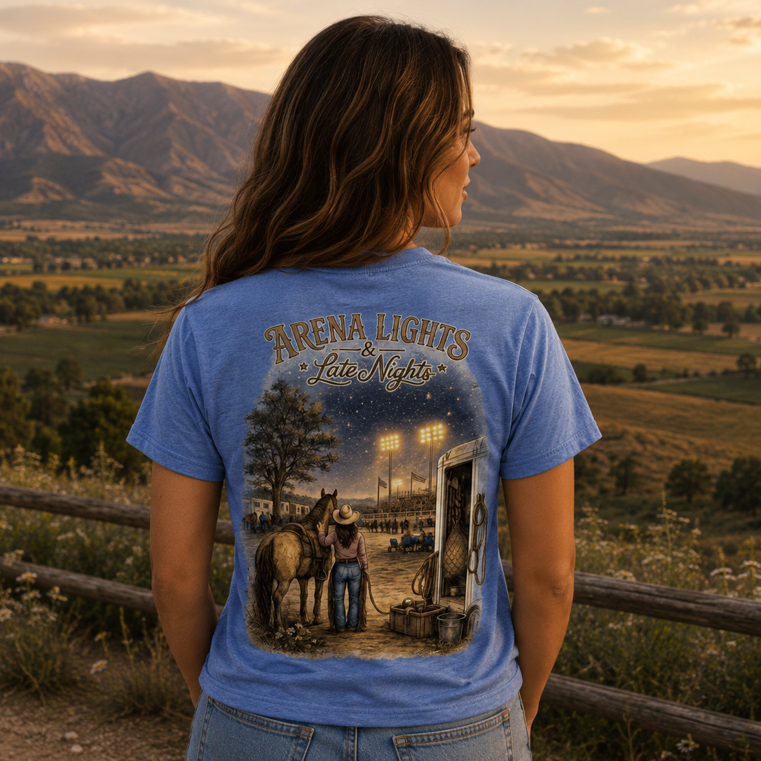 A woman stands outdoors in a scenic mountain valley at sunset, viewed from behind, wearing a light blue graphic T-shirt with the text 'Arena Lights Late Night' and a western-themed illustration on the back.