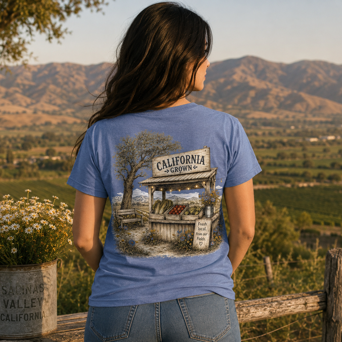 A woman stands outdoors in a rural landscape, viewed from behind, wearing a blue graphic T-shirt with a large printed illustration of a California orchard stand and mountains. The shirt is the product being modeled in a real environment, so this is Lifestyle rather than Product/Packaging.