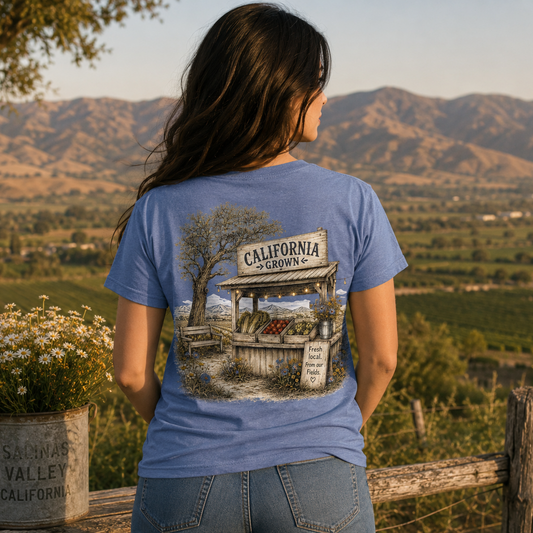 A woman stands outdoors in a rural landscape, viewed from behind, wearing a blue graphic T-shirt with a large printed illustration of a California orchard stand and mountains. The shirt is the product being modeled in a real environment, so this is Lifestyle rather than Product/Packaging.