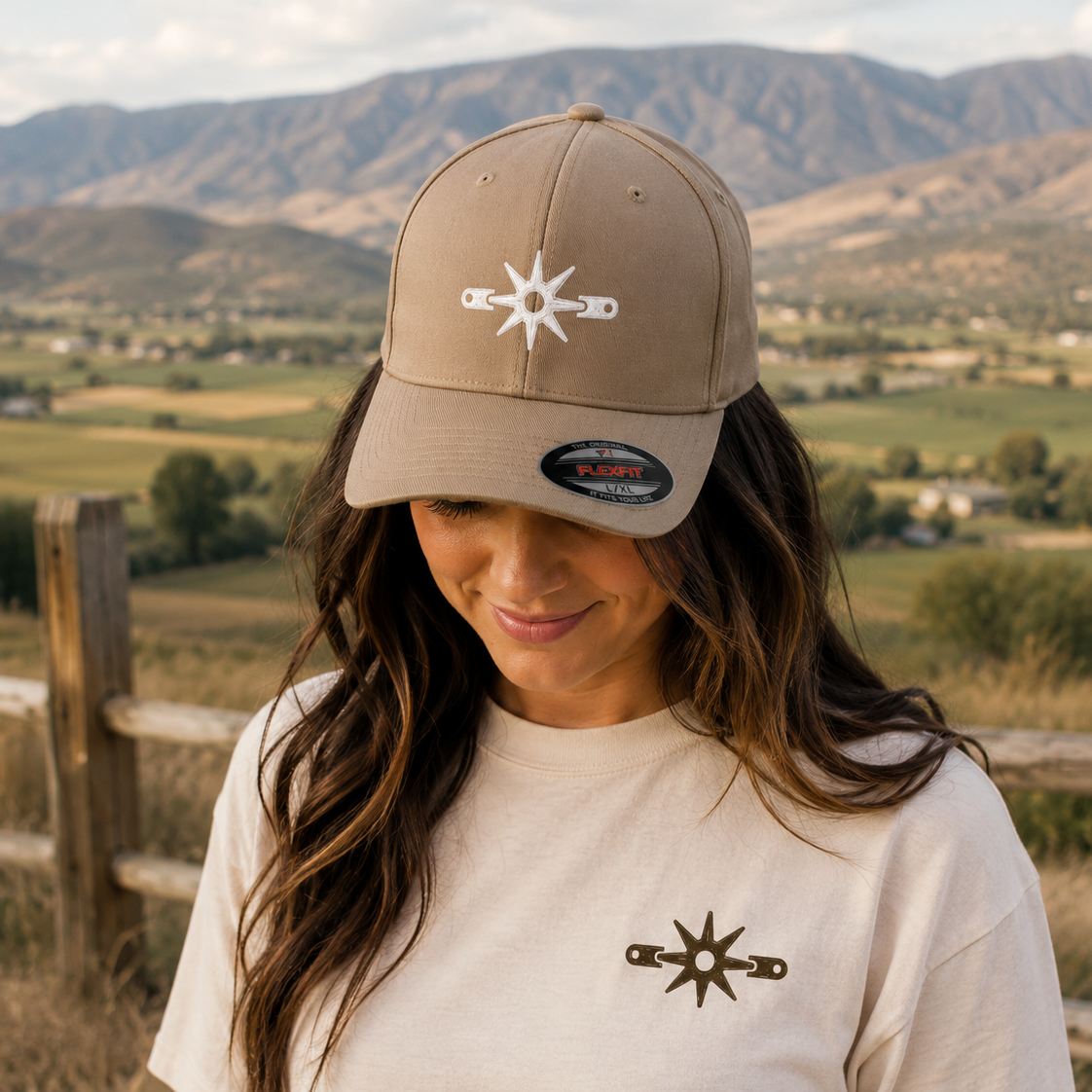 A woman wearing a tan baseball cap and a cream t-shirt, both featuring a white and brown compass star logo, set against a blurred mountain landscape.