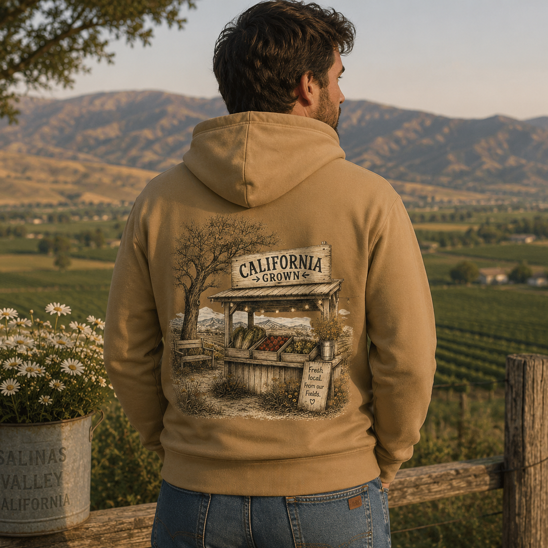 A man wearing a tan hoodie with a large 'California Grown' graphic on the back, standing outdoors in front of a scenic vineyard landscape.