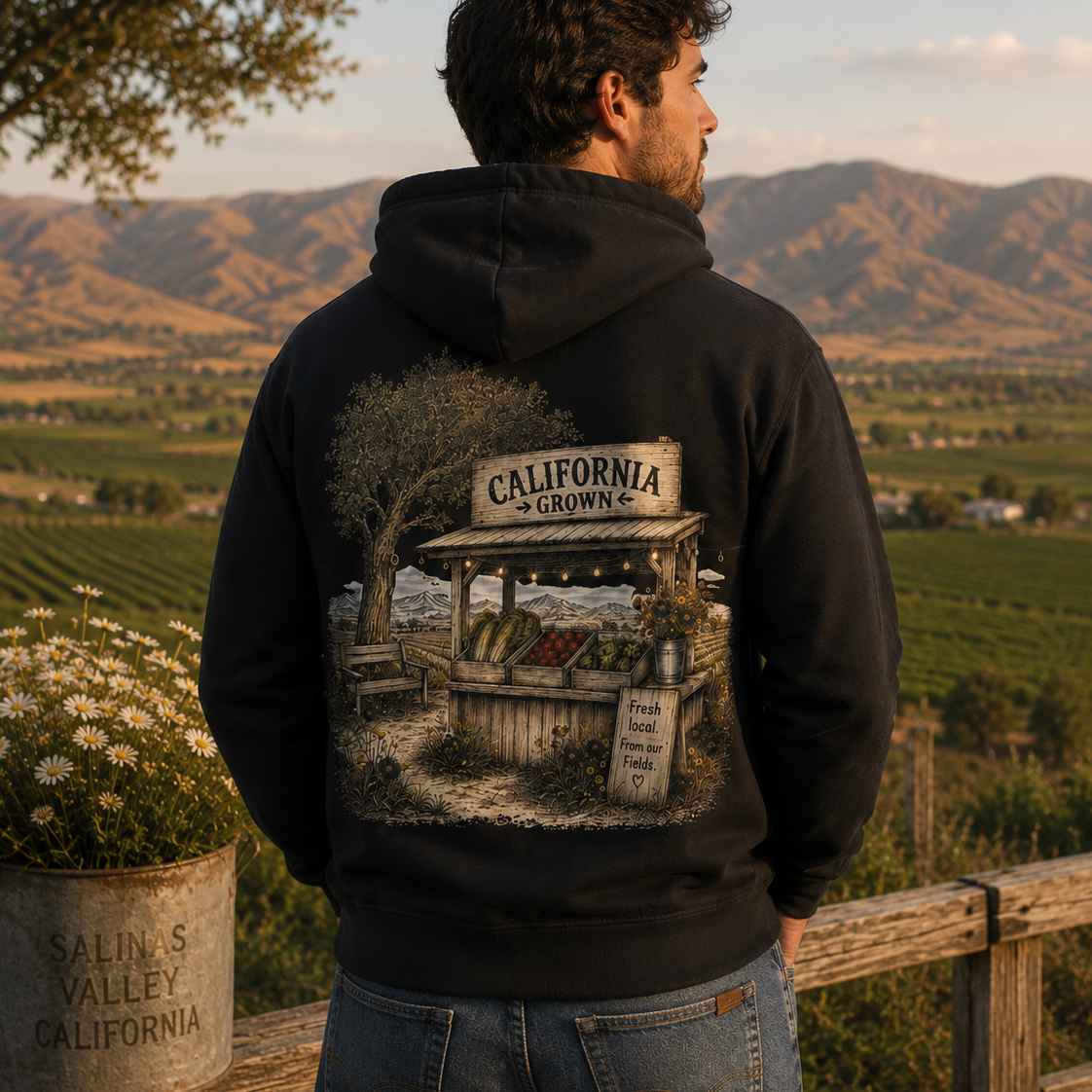 A man wearing a black hoodie with a large back graphic of a California wine cellar scene, standing outdoors in front of a vineyard landscape at sunset.