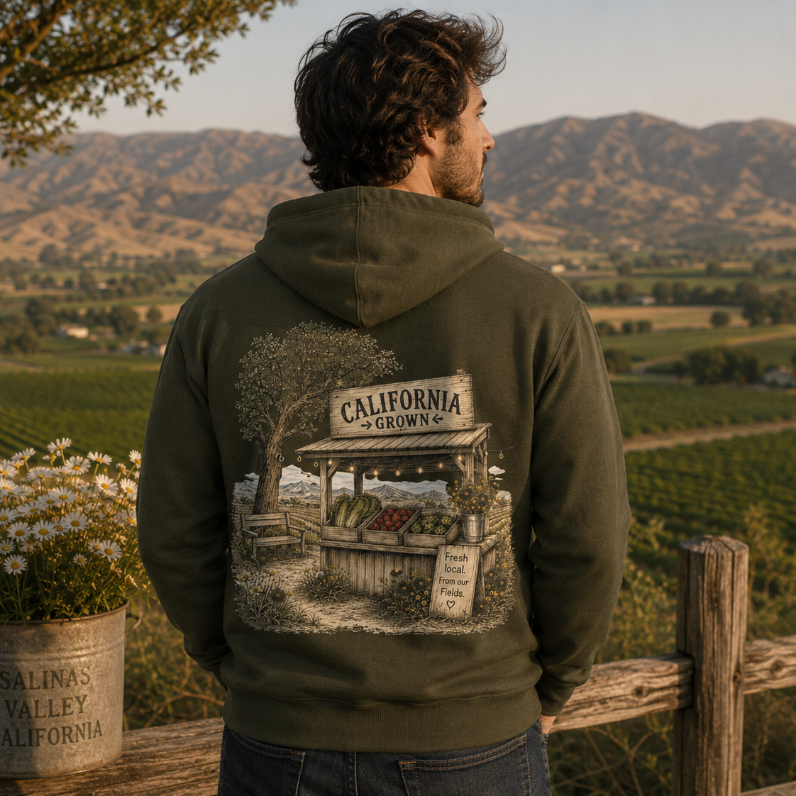 A man stands outdoors in a rural landscape wearing an olive green hoodie, viewed from behind. The hoodie features a large graphic print on the back depicting a California vineyard scene with the text 'CALIFORNIA GROWN'.