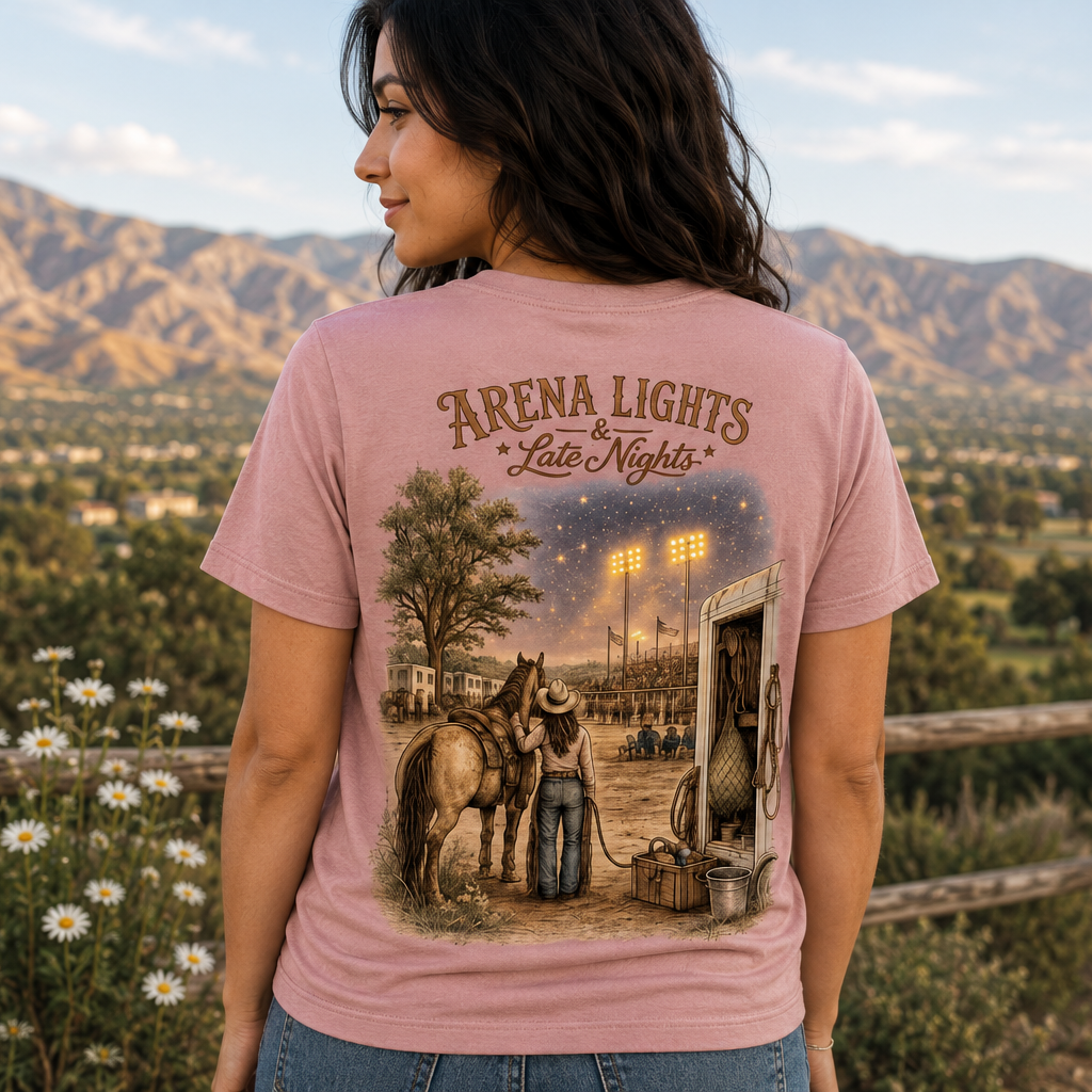 A woman wearing a light pink graphic t-shirt, viewed from behind, with a scenic outdoor background of mountains, a wooden fence, and wildflowers. The t-shirt features a large graphic print of a western night scene with a cowboy, a horse, and a gas station, with the text 'ARENA LIGHTS Late Nights'.